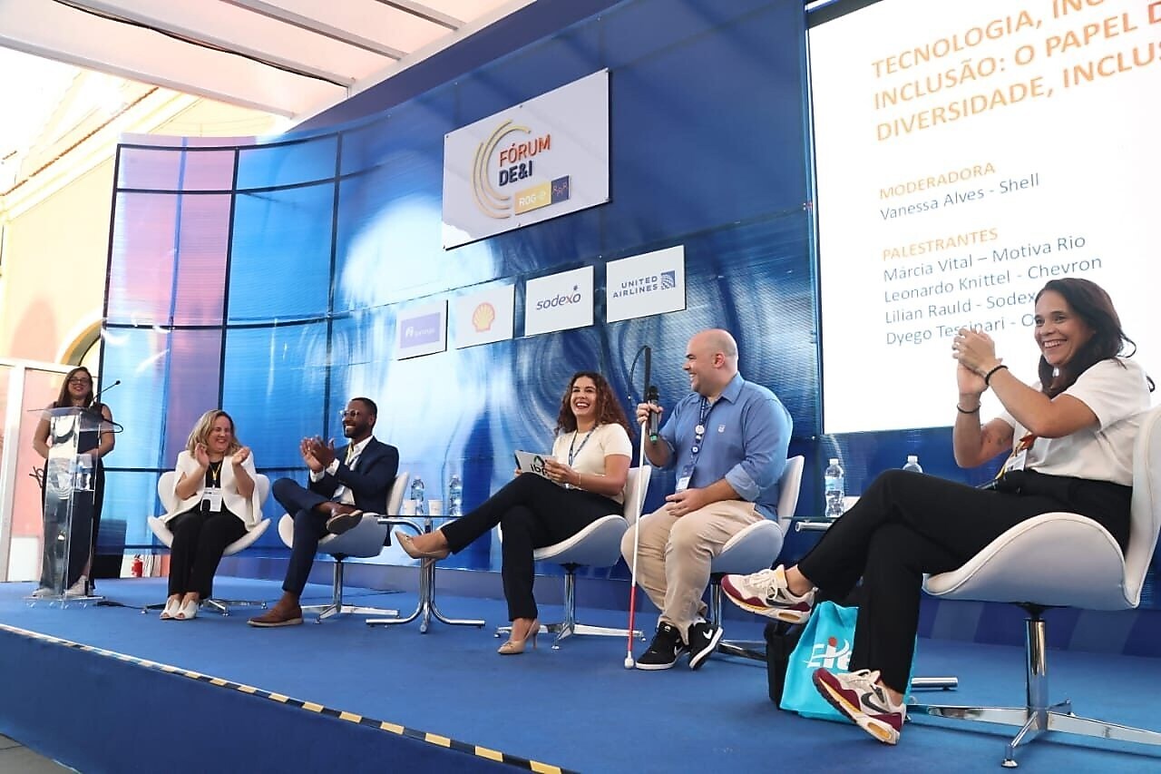 Cinco palestrantes, incluindo quatro mulheres e um homem, estão sentados em cadeiras brancas no palco durante um painel de discussão sobre diversidade e inclusão. Um banner ao fundo exibe o nome do evento: 'Fórum DE&I', com logotipos de patrocinadores como Shell, Sodexo e United Airlines. À direita, uma tela destaca o tema da sessão, focado em tecnologia e inclusão, com os nomes dos participantes. Todos parecem engajados, sorrindo e aplaudindo.