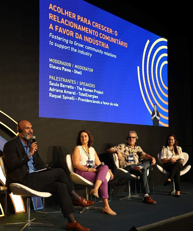 A imagem mostra quatro palestrantes sentados em cadeiras de um painel de discussão. À esquerda, o moderador Glauco Paiva, da Shell, está segurando um microfone enquanto fala, vestindo um terno escuro e uma camisa azul. Ao lado dele, três palestrantes estão sentados, incluindo Adriana Amaral, da TotalEnergies, vestindo calça roxa e blusa clara, e outros dois palestrantes, um homem de camisa estampada e uma mulher de calça preta e blusa branca. Ao fundo, um grande telão azul exibe o título da palestra: “Acolher para crescer: o relacionamento comunitário a favor da indústria” e os nomes dos participantes.