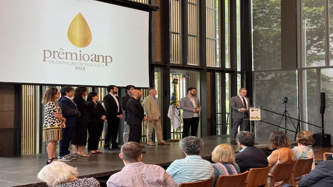 Cerimônia do Prêmio ANP de Inovação Tecnológica 2024. Um grupo de pessoas está no palco, com dois palestrantes ao centro, um deles segurando um troféu. O logo do evento está projetado ao fundo. Plateia acompanha a premiação.