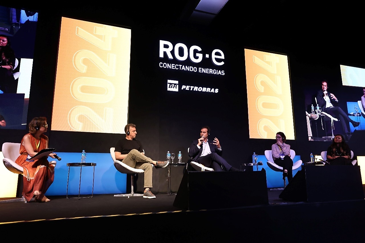 Painel de discussão no evento ROG.e 2024, com cinco palestrantes sentados em cadeiras brancas no palco, discutindo o tema 'Conectando Energias'. Dois grandes painéis digitais atrás dos participantes exibem o logotipo do evento, a marca Petrobras, e o número 2024 em destaque. Todos os participantes estão usando fones de ouvido, e há garrafas de água nas mesas laterais.