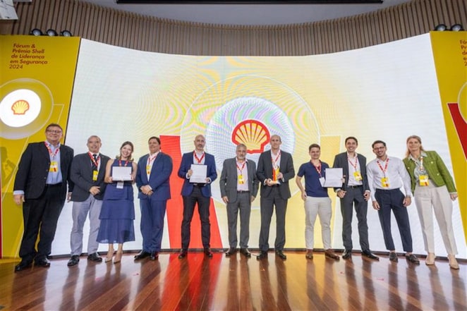 Grupo de nove pessoas em um palco, segurando certificados e troféus durante o Fórum e Prêmio Shell de Liderança em Segurança 2024. Ao fundo, um telão exibe o logotipo da Shell. Todos estão vestindo crachás e sorrindo para a câmera. Nas laterais, banners amarelos com o logotipo da Shell e o título do evento.