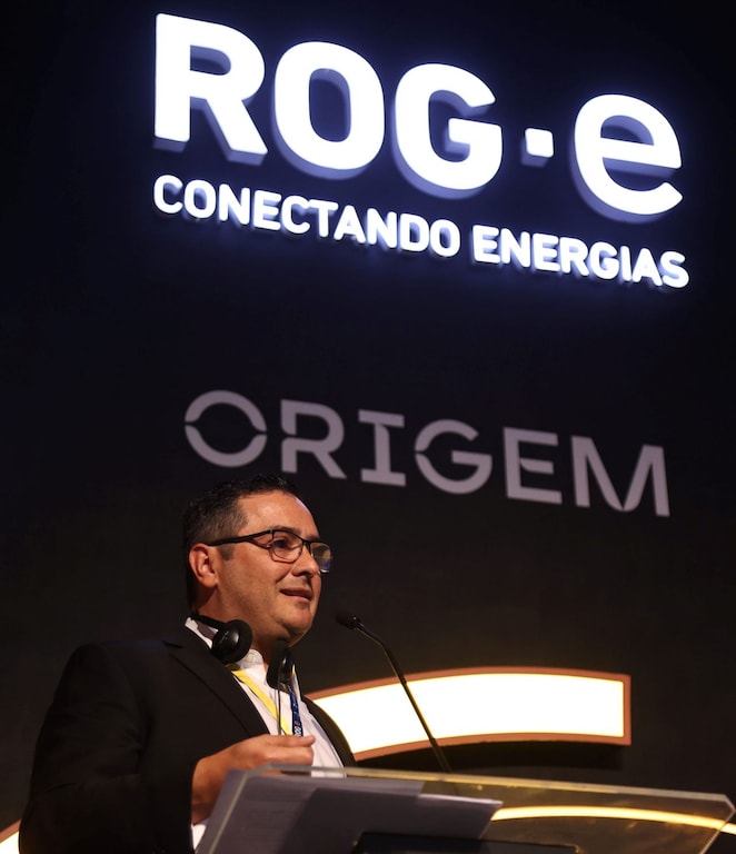 Homem de óculos, usando terno preto e camisa branca, fala em um púlpito durante um evento. No fundo, há um grande letreiro iluminado com as palavras 'ROG·e Conectando Energias' e 'ORIGEM'. Ele está segurando um controle e tem um fone de ouvido ao redor do pescoço.