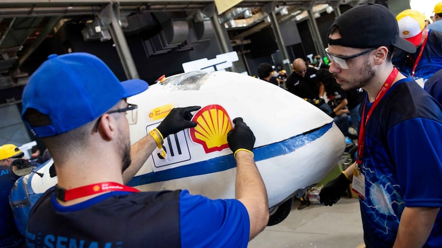Uma equipe de pessoas vestindo camisetas azuis com o logotipo do SENAI nas costas está trabalhando em um grande modelo de veículo branco e azul. A pessoa em primeiro plano está colocando o logotipo da Shell no veículo. O ambiente parece ser um evento ou área de competição interna, sugerido pela estrutura do local e outros participantes ao fundo usando bonés amarelos.