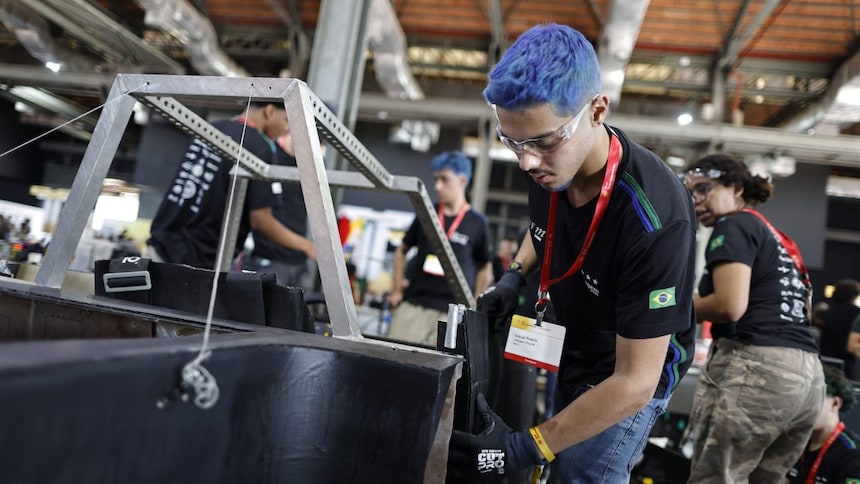 Um grupo de pessoas em um ambiente interno, que parece ser um evento de tecnologia ou jogos. O foco está em uma pessoa com cabelo azul, em primeiro plano, vestindo uma camiseta azul marinho e um crachá no pescoço. Há outras pessoas ao fundo. O ambiente inclui vários cabos e dispositivos eletrônicos, sugerindo que pode ser uma competição, oficina ou evento colaborativo relacionado à tecnologia ou computação.