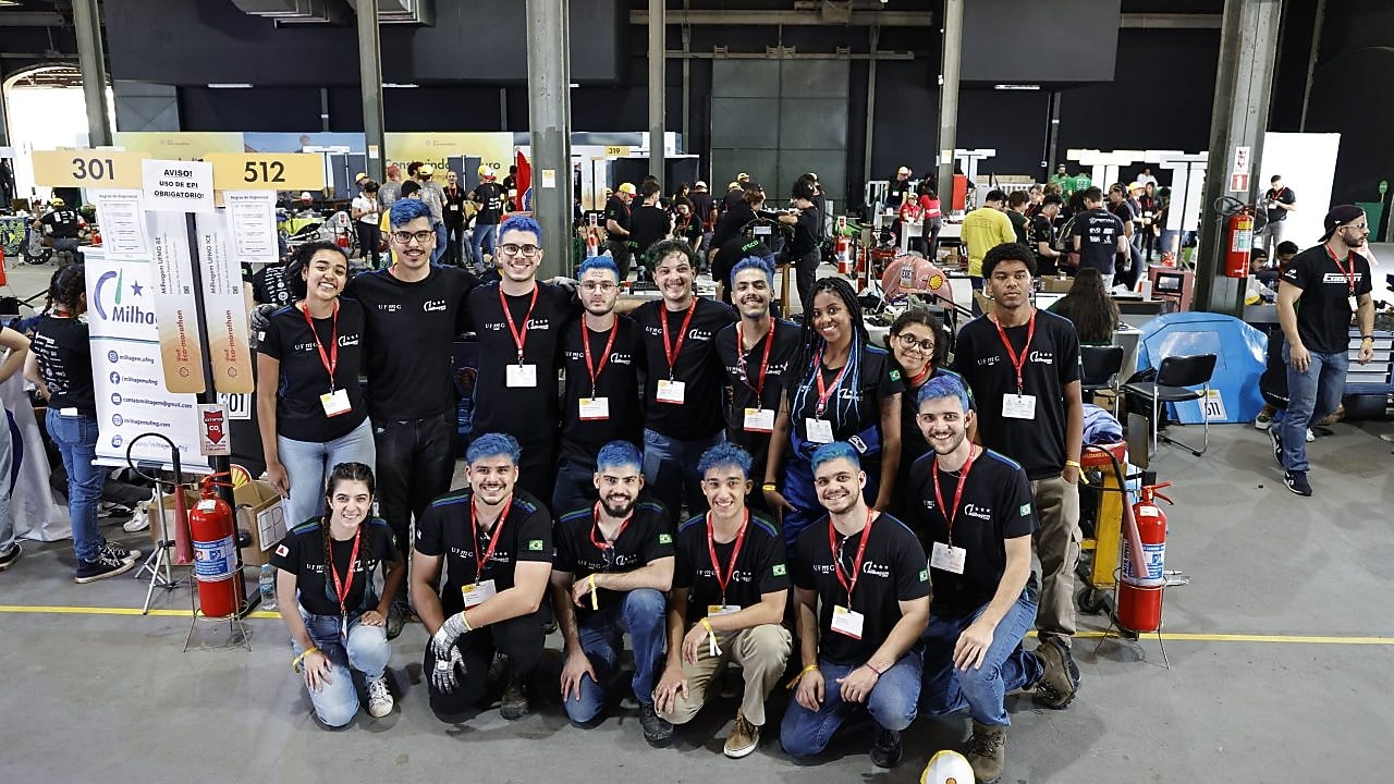 Um grupo de pessoas em um ambiente interno, possivelmente em um evento de robótica ou engenharia. As pessoas estão todas vestidas casualmente, com camiseta azul marinho e calças jeans (na maioria) e usando crachás, sugerindo que são participantes ou visitantes. Ao fundo, há banners com números e outros grupos de pessoas, indicando que pode ser uma competição ou exposição.