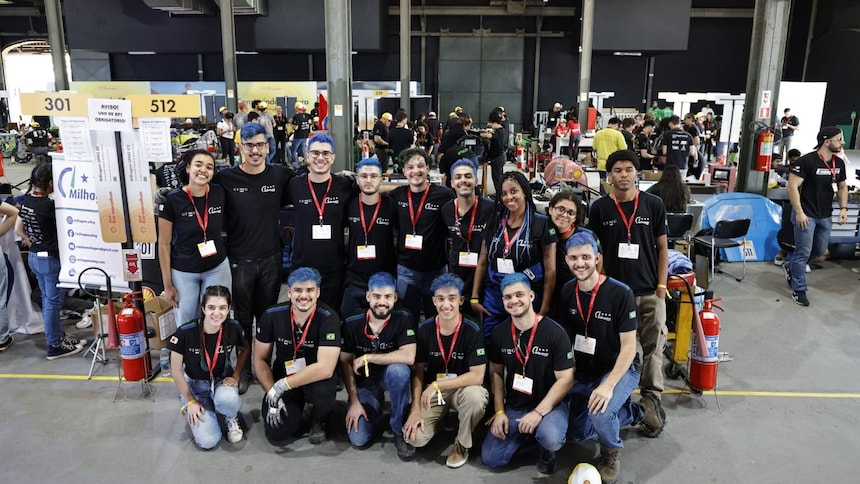 Um grupo de pessoas em um ambiente interno, possivelmente em um evento de robótica ou engenharia. As pessoas estão todas vestidas casualmente, com camiseta azul marinho e calças jeans (na maioria) e usando crachás, sugerindo que são participantes ou visitantes. Ao fundo, há banners com números e outros grupos de pessoas, indicando que pode ser uma competição ou exposição.