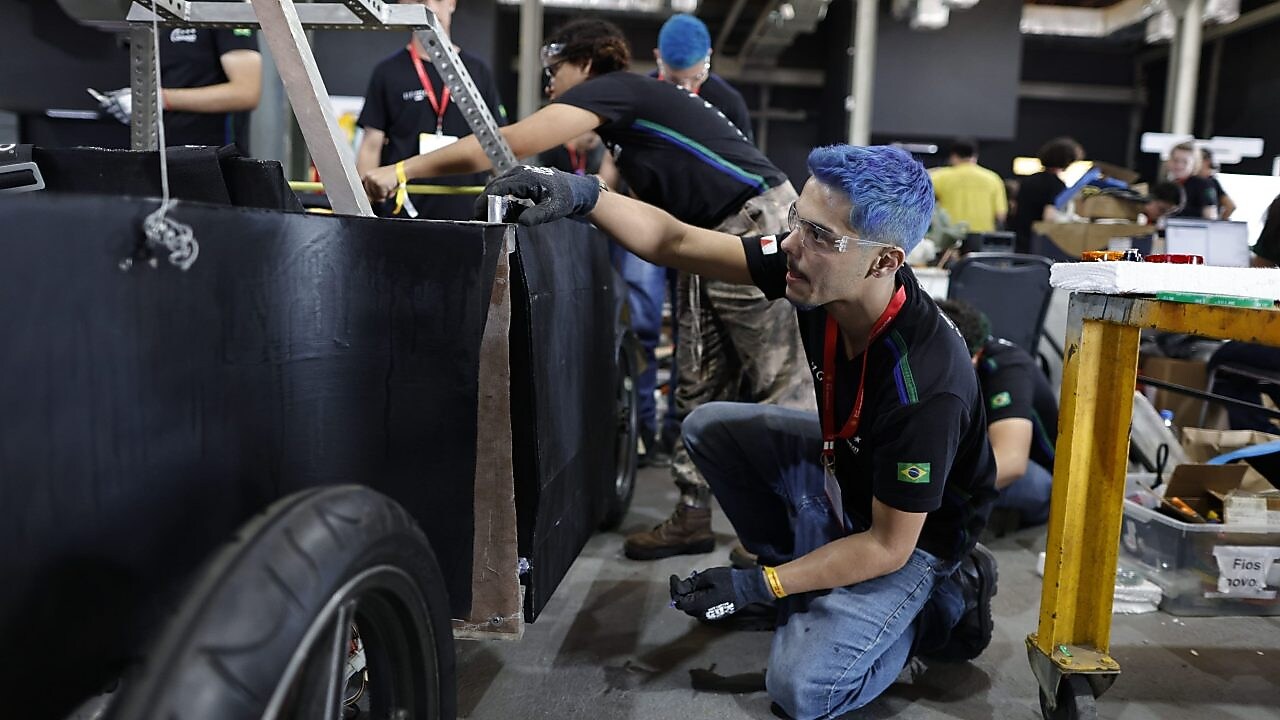 Um grupo de pessoas trabalhando em uma grande estrutura com rodas ou veículo. Uma pessoa está ajoelhada em primeiro plano, usando luvas e focada em uma tarefa ao lado da estrutura. Ao fundo, outras pessoas estão envolvidas em várias atividades ao redor da estrutura e equipamentos. O ambiente sugere uma oficina ou local de construção, com ferramentas e materiais visíveis, indicando trabalho em equipe e manual em um projeto que requer habilidades técnicas e labor físico.
