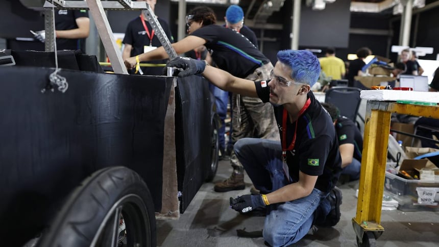 Um grupo de pessoas trabalhando em uma grande estrutura com rodas ou veículo. Uma pessoa está ajoelhada em primeiro plano, usando luvas e focada em uma tarefa ao lado da estrutura. Ao fundo, outras pessoas estão envolvidas em várias atividades ao redor da estrutura e equipamentos. O ambiente sugere uma oficina ou local de construção, com ferramentas e materiais visíveis, indicando trabalho em equipe e manual em um projeto que requer habilidades técnicas e labor físico.