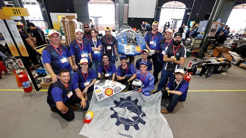 Um grupo de pessoas em pé atrás de um pequeno veículo aerodinâmico com o número 311. As pessoas estão vestindo camisetas azuis combinando e crachás pendendo do pescoço. Eles estão em um grande espaço interno que parece ser uma área de exposição ou oficina, sugerido pela presença de outros projetos e banners ao fundo. Um grande banner com um logotipo está estendido na frente do grupo no chão, e uma pessoa está segurando um capacete branco. 