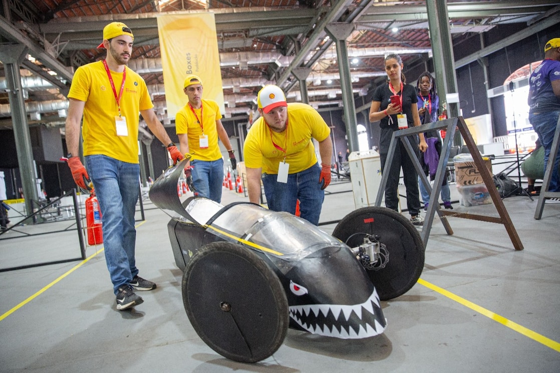 Grupo de pessoas vestindo camisetas amarelas e calças jeans ao redor de um veículo preto aerodinâmico com design de tubarão, incluindo dentes brancos e olhos vermelhos pintados na frente. O veículo tem grandes rodas pretas e está em um ambiente interno com estruturas industriais ao fundo. Todos estão usando crachás de identificação em cordões e alguns estão usando bonés.