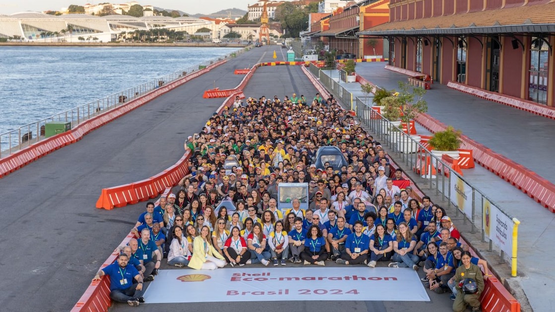 A imagem mostra um grande grupo de pessoas reunidas para uma fotografia na Shell Eco-marathon Brasil 2024. Os participantes estão ao ar livre, com alguns sentados e outros em pé ao redor de uma faixa larga que exibe o nome do evento. O cenário é uma área espaçosa com uma pista no Pier Mauá, no Rio de Janeiro, com edifícios e colinas visíveis ao fundo sob um céu claro. A imagem captura o espírito coletivo dos participantes em uma competição automotiva ecológica, destacando sua unidade e entusiasmo pela inovação sustentável.
