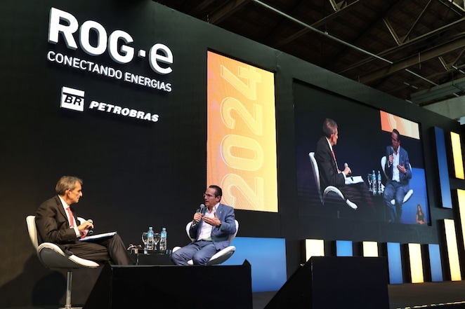Dois executivos participam de uma conversa em um palco no evento ROG.e 2024, com o tema 'Conectando Energias'. O fundo do palco exibe logotipos da Petrobras e a data '2024'. Um dos executivos segura um microfone enquanto gesticula, e o outro faz anotações. Ambos estão sentados em cadeiras modernas, com garrafas de água e copos sobre a mesa entre eles. Ao lado, um telão exibe o evento em tempo real.