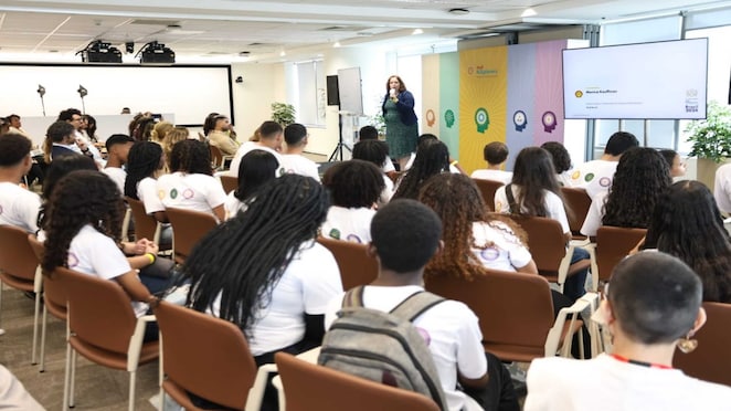 Auditório com jovens usando camisetas brancas sentados em cadeiras, assistindo a uma palestra de uma mulher em pé ao lado de um púlpito e um telão. O ambiente é moderno, com painel colorido ao fundo.
