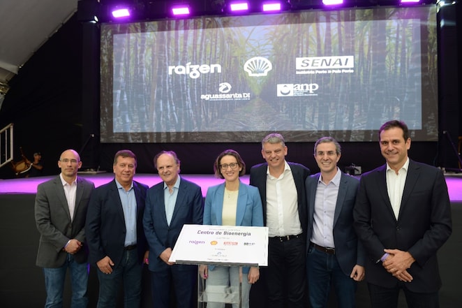 Grupo de sete pessoas, incluindo homens e uma mulher, posam sorrindo para foto durante um evento de inauguração. Ao fundo, um telão exibe as marcas Raízen, Shell, SENAI e ANP, com uma imagem de fundo que remete a plantações de cana-de-açúcar. O grupo está ao redor de uma placa que anuncia o "Centro de Bioenergia". A iluminação do palco é predominante em tons de roxo e as pessoas estão vestidas formalmente, com trajes sociais.