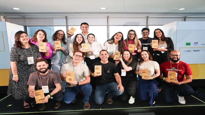 Grupo de pessoas segurando troféus de madeira, sorrindo em frente a um painel com logotipos de um evento de premiação