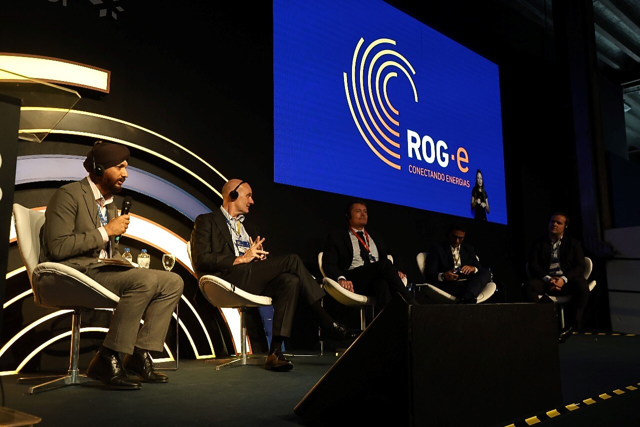 Painel com cinco pessoas sentadas no palco do evento ROG.e, que foca na conexão de energias. Um dos palestrantes, usando um terno cinza e um turbante, segura um microfone enquanto fala. Os outros participantes do painel, vestidos com trajes formais, escutam atentamente. Atrás deles, uma grande tela exibe o logo do evento em azul e laranja. O palco tem iluminação moderna e a plateia não está visível.