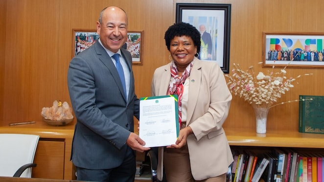 Dois indivíduos em pé em um ambiente de escritório, sorrindo e segurando juntos um documento. O homem à esquerda está vestindo terno e gravata, enquanto a mulher à direita veste um terno bege com um lenço no pescoço. Ao fundo, há painéis de madeira, estantes com livros e fotografias emolduradas.