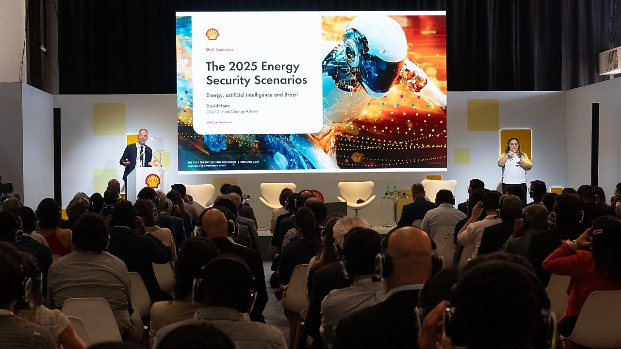 Auditório cheio de pessoas assistindo a uma apresentação da Shell sobre segurança energética em 2025. No palco, um palestrante em pé ao lado de um púlpito fala diante de um telão que exibe o título 'The 2025 Energy Security Scenarios – Energy, artificial intelligence and Brazil', com a imagem de um robô e elementos digitais futuristas. À direita, uma intérprete de Libras traduz a fala. O público, de costas, usa fones de ouvido para acompanhar a apresentação.