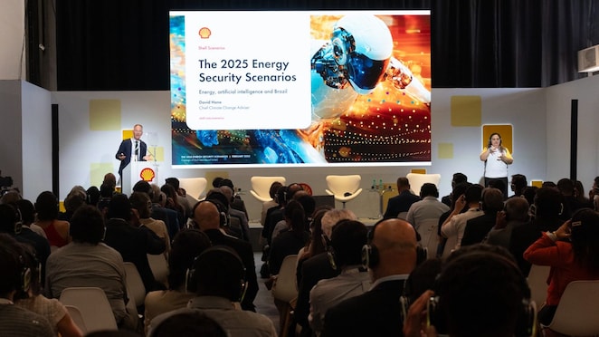 Auditório cheio de pessoas assistindo a uma apresentação da Shell sobre segurança energética em 2025. No palco, um palestrante em pé ao lado de um púlpito fala diante de um telão que exibe o título 'The 2025 Energy Security Scenarios – Energy, artificial intelligence and Brazil', com a imagem de um robô e elementos digitais futuristas. À direita, uma intérprete de Libras traduz a fala. O público, de costas, usa fones de ouvido para acompanhar a apresentação.
