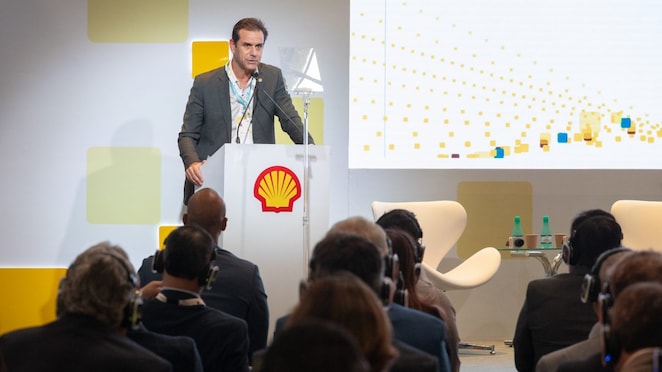 Homem de terno cinza fala em um púlpito com o logotipo da Shell, durante uma conferência. Ele segura o microfone enquanto olha para o público, que aparece de costas usando fones de ouvido. Ao lado do palco, há cadeiras brancas e garrafas de água sobre uma mesa de vidro. No telão ao fundo, aparecem elementos gráficos em forma de quadrados amarelos e azuis.
