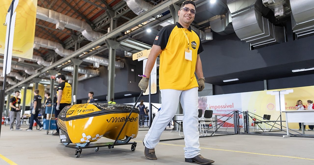 Jovem com crachá e luvas de proteção trabalha no interior de um protótipo de veículo durante a Shell Eco-marathon. Ele sorri enquanto ajusta componentes, cercado por outros integrantes da equipe em um espaço interno de competição