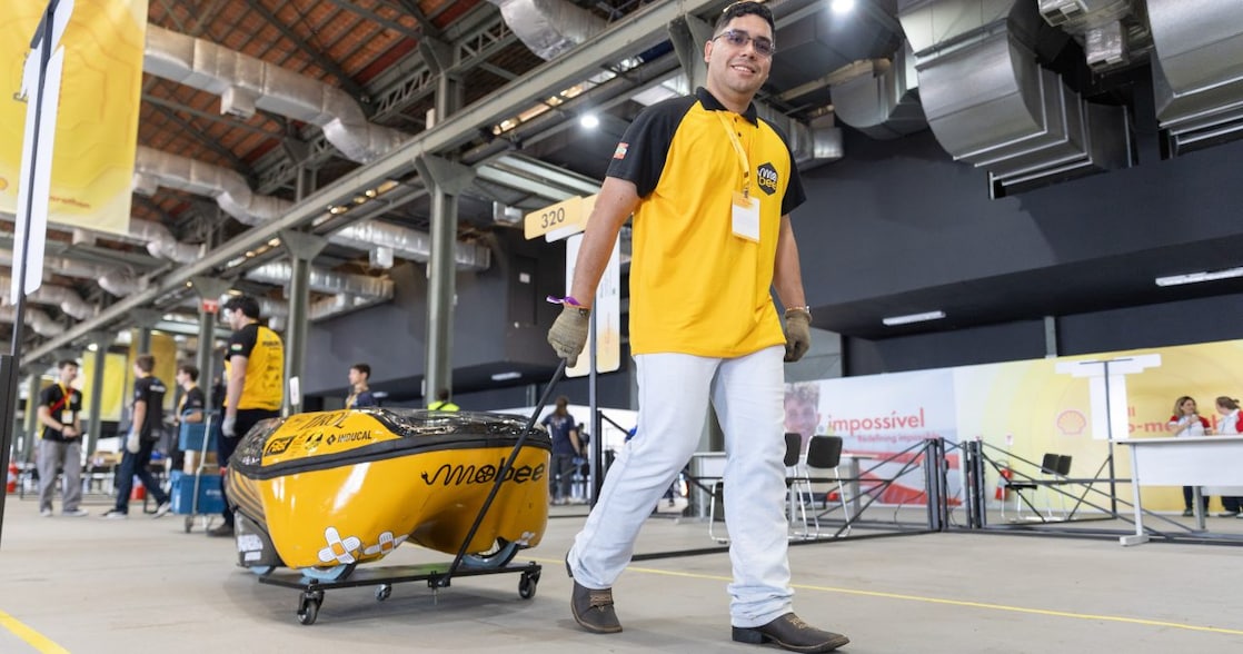 Jovem com crachá e luvas de proteção trabalha no interior de um protótipo de veículo durante a Shell Eco-marathon. Ele sorri enquanto ajusta componentes, cercado por outros integrantes da equipe em um espaço interno de competição