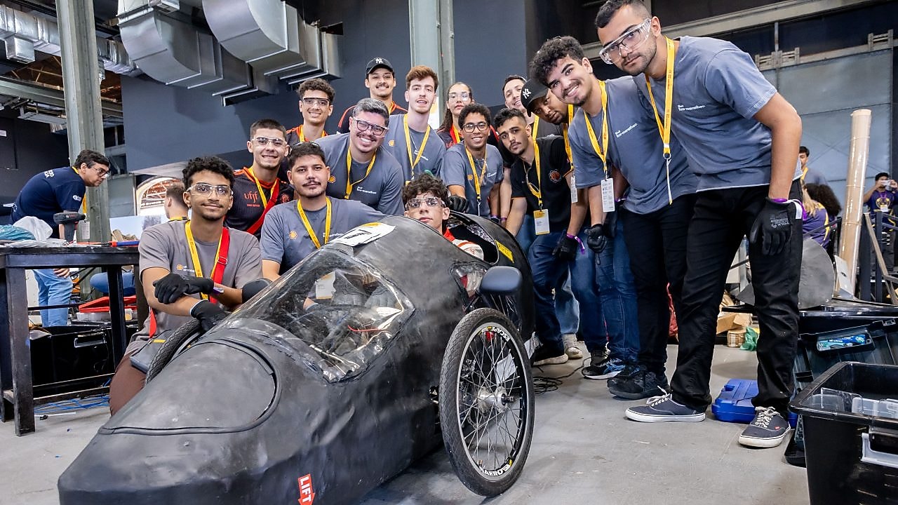 Grupo de estudantes de engenharia posa sorridente em torno de um protótipo de carro elétrico aerodinâmico, de três rodas, construído para competição de eficiência energética.