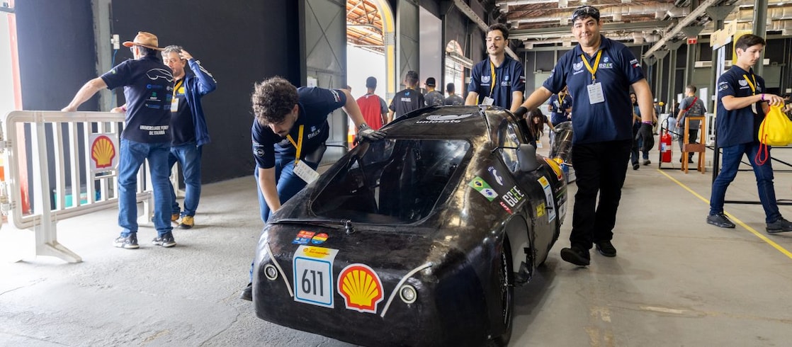 Estudante pilota um carro protótipo futurista de eficiência energética durante a Shell Eco-marathon. O veículo de número 314, em azul e branco, traz o logotipo da Shell na frente e está em movimento na pista de competição.