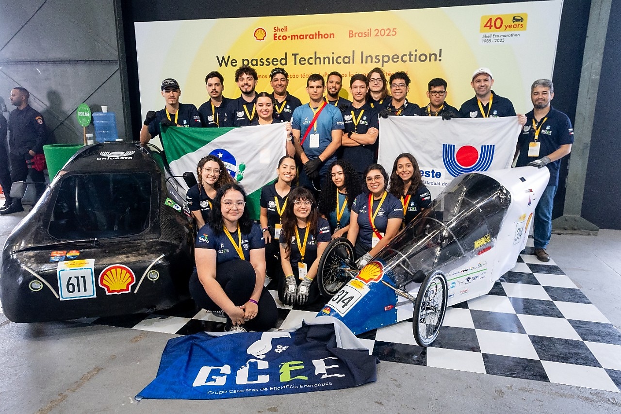 Equipe de estudantes posa sorridente após aprovação na inspeção técnica da Shell Eco-marathon Brasil 2025. O grupo segura bandeiras do Brasil, da Unioeste e do GCEE, posicionados ao lado de dois carros protótipos de eficiência energética, numerados 611 e 314, com o logotipo da Shell na frente. Ao fundo, banner com a frase 'We passed Technical Inspection!' e a marca dos 40 anos da competição.