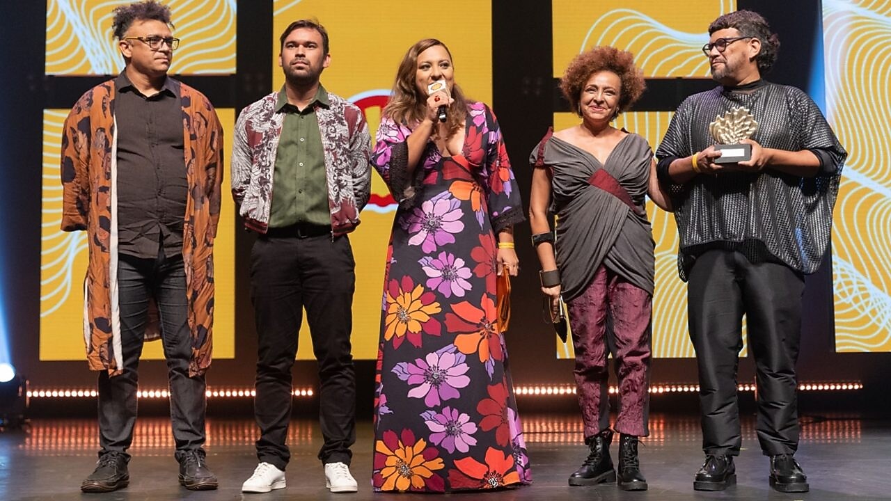 Cinco pessoas estão em um palco iluminado, em frente a um telão amarelo com padrões brancos e pretos. Da esquerda para a direita: um homem de óculos e cabelo grisalho curto usa camisa preta, calça jeans e um kimono marrom estampado; um homem de barba curta veste camisa verde, calça preta e jaqueta estampada; uma mulher no centro segura um microfone e usa vestido longo preto com grandes flores roxas, lilases, vermelhas, laranjas e amarelas; uma mulher com cabelo crespo curto veste blusa cinza transpassada com detalhe bordô, calça roxa e botas pretas; um homem segura um troféu dourado em forma de concha, veste blusa preta larga e calça preta.