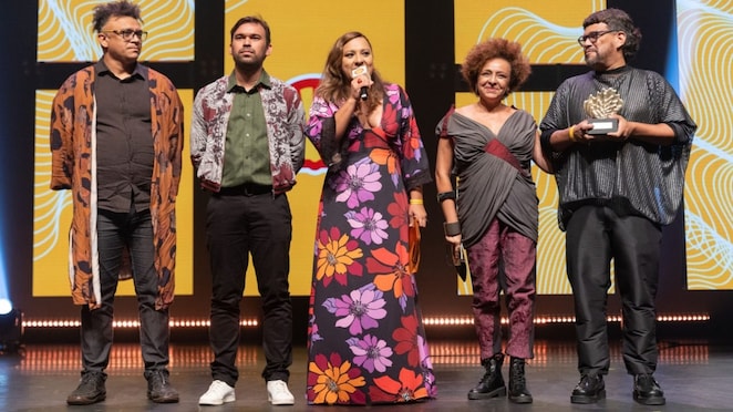 Cinco pessoas estão em um palco iluminado, em frente a um telão amarelo com padrões brancos e pretos. Da esquerda para a direita: um homem de óculos e cabelo grisalho curto usa camisa preta, calça jeans e um kimono marrom estampado; um homem de barba curta veste camisa verde, calça preta e jaqueta estampada; uma mulher no centro segura um microfone e usa vestido longo preto com grandes flores roxas, lilases, vermelhas, laranjas e amarelas; uma mulher com cabelo crespo curto veste blusa cinza transpassada com detalhe bordô, calça roxa e botas pretas; um homem segura um troféu dourado em forma de concha, veste blusa preta larga e calça preta.