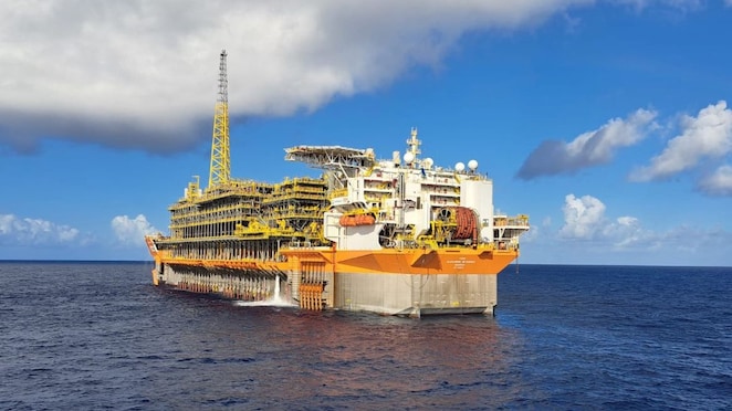 Plataforma flutuante de produção de petróleo (FPSO), de grande porte, com estrutura predominantemente amarela e branca, flutuando em alto-mar sob céu azul com algumas nuvens. A embarcação, equipada com módulos industriais e torre alta, está parcialmente descarregando água pelo casco.