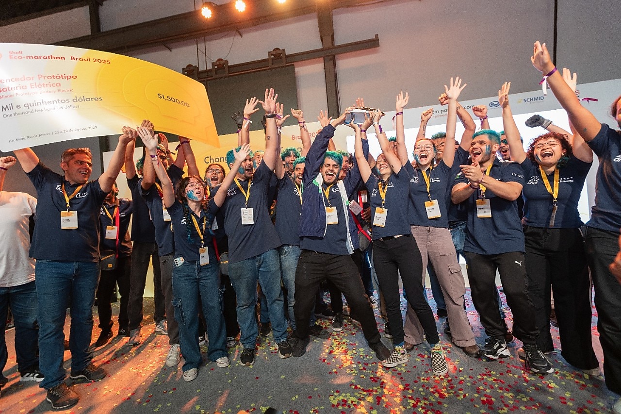 Grupo de estudantes comemorando vitória na Shell Eco-marathon Brasil 2025. Eles vestem camisetas azuis e crachás, levantam os braços em celebração e seguram um grande cheque amarelo de 1.500 dólares, rodeados por confetes coloridos no chão.