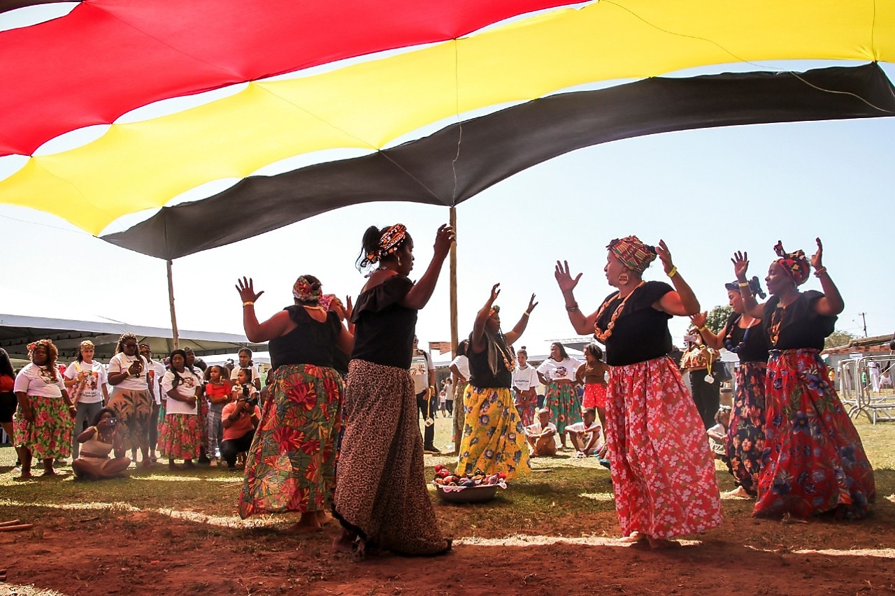 Grupo de mulheres negras dança em círculo ao ar livre, usando saias coloridas e turbantes, sob uma grande faixa de tecido nas cores vermelho, amarelo e preto. Ao fundo, outras pessoas observam e participam da celebração em um ambiente ensolarado. 