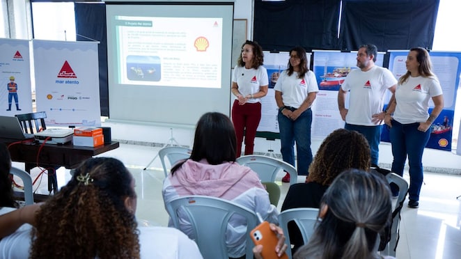 Apresentação do Projeto Mar Atento, com quatro integrantes de camiseta branca falando para um grupo de participantes sentados. Ao fundo, há um telão com informações sobre o projeto e banners com os logotipos da Shell e do Mar Atento em um ambiente de evento educativo.