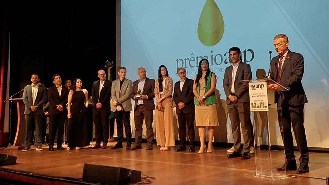 A imagem mostra um palco de auditório durante a cerimônia do Prêmio ANP de Inovação Tecnológica. Cerca de doze pessoas estão alinhadas em fileira, vestidas de maneira formal, segurando troféus ou aguardando a fala oficial. À direita, um homem apresenta o evento em um púlpito com a marca da ANP e do Ministério de Minas e Energia. Ao fundo, o telão exibe o símbolo do prêmio, uma gota dourada sobre fundo claro, iluminando a cena com caráter solene.