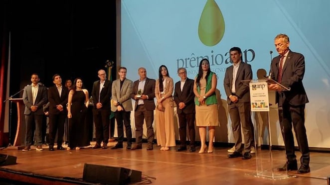 A imagem mostra um palco de auditório durante a cerimônia do Prêmio ANP de Inovação Tecnológica. Cerca de doze pessoas estão alinhadas em fileira, vestidas de maneira formal, segurando troféus ou aguardando a fala oficial. À direita, um homem apresenta o evento em um púlpito com a marca da ANP e do Ministério de Minas e Energia. Ao fundo, o telão exibe o símbolo do prêmio, uma gota dourada sobre fundo claro, iluminando a cena com caráter solene.