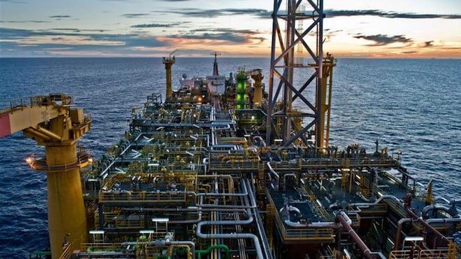 Visão aérea de uma plataforma marítima de petróleo ao entardecer, com inúmeras tubulações, estruturas metálicas e equipamentos industriais, cercada pelo oceano e iluminada pela luz do pôr do sol.