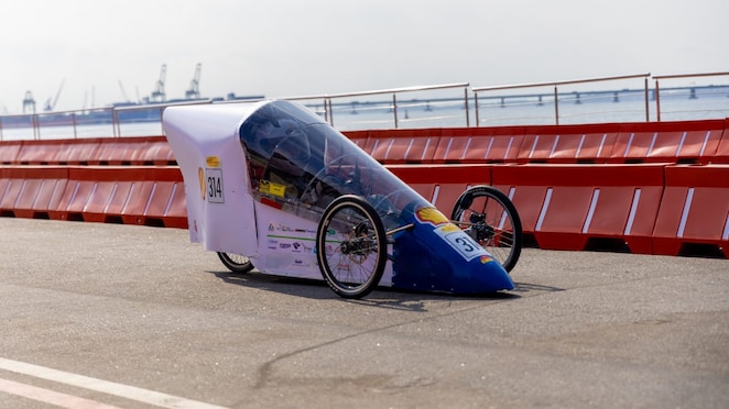 Veículo protótipo de eficiência energética de três rodas, com carroceria aerodinâmica roxa e azul, participando da competição Shell Eco-marathon em uma pista próxima ao mar.