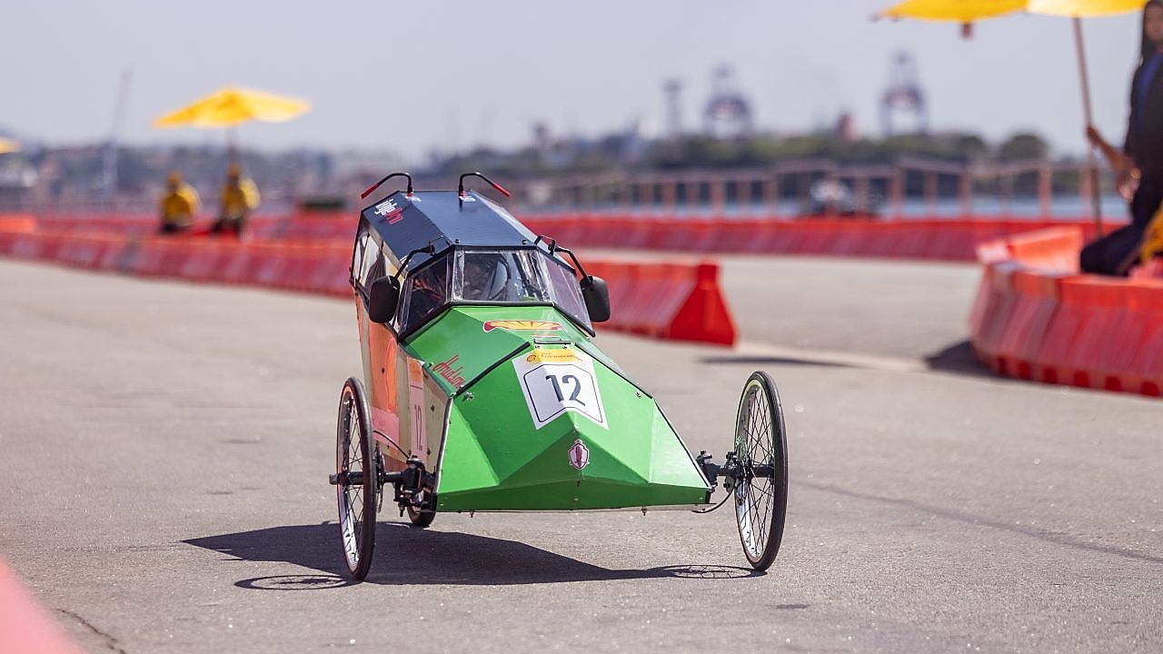 Carro protótipo verde e preto de três rodas, número 12, participando de uma competição de eficiência energética em pista delimitada por barreiras laranjas, em um dia ensolarado.