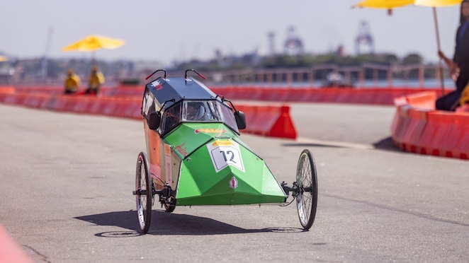 Carro protótipo verde e preto de três rodas, número 12, participando de uma competição de eficiência energética em pista delimitada por barreiras laranjas, em um dia ensolarado.