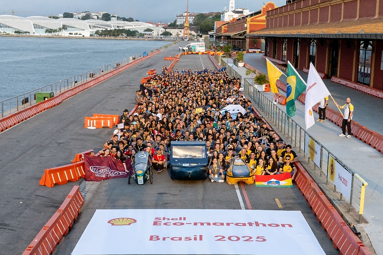 Grupo de participantes da Shell Eco-marathon Brasil 2025 posando atrás de três protótipos de veículos sustentáveis, com bandeiras e faixas de equipes, em frente ao Museu do Amanhã, no Rio de Janeiro.