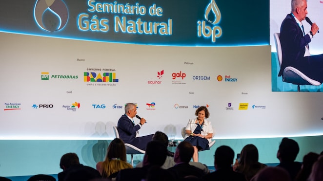 Duas pessoas participam de um painel no palco do Seminário de Gás Natural, organizado pelo Instituto Brasileiro de Petróleo e Gás (IBP). Elas estão sentadas em cadeiras brancas, conversando com microfones em mãos, diante de uma plateia. Ao fundo, há um painel com o logotipo do evento e de patrocinadores como Petrobras, Shell Energy, Governo Federal, Galp, Equinor, Eneva e outros.