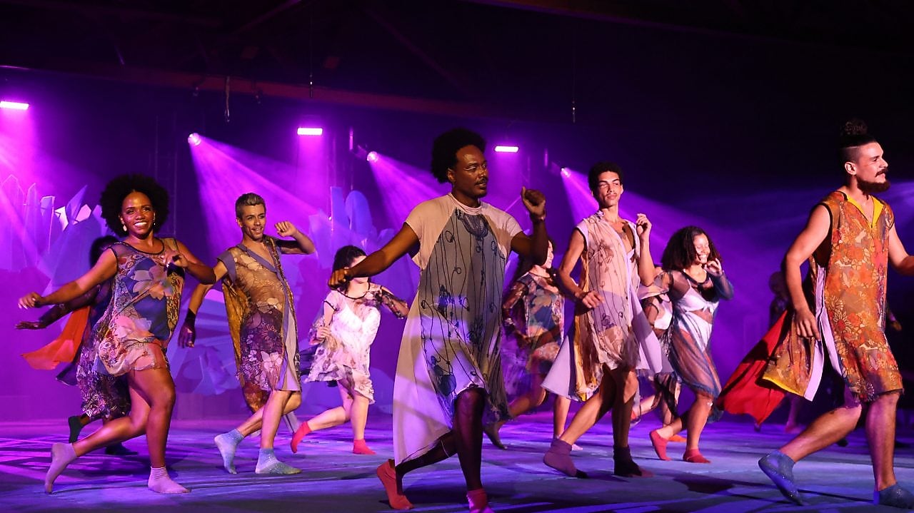 Grupo de dançarinos se apresenta em um palco iluminado por luzes roxas, usando trajes coloridos e fluidos. Eles se movem de forma sincronizada, transmitindo energia e alegria