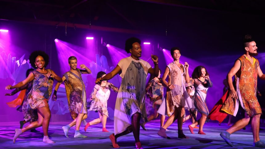 Grupo de dançarinos se apresenta em um palco iluminado por luzes roxas, usando trajes coloridos e fluidos. Eles se movem de forma sincronizada, transmitindo energia e alegria