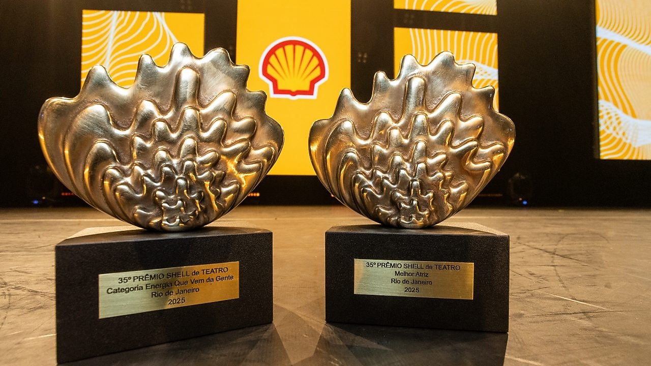 Dois troféus do 35º Prêmio Shell de Teatro estão posicionados lado a lado sobre o palco. As estatuetas metálicas têm o formato de uma concha estilizada e repousam sobre bases pretas com placas douradas. A placa da estatueta à esquerda indica a categoria "Energia Que Vem da Gente", e a da direita, "Melhor Atriz", ambas referentes à edição de 2025 no Rio de Janeiro. Ao fundo, o palco está decorado com painéis amarelos e o logotipo da Shell em destaque.