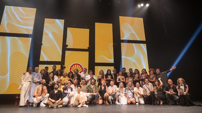 Grupo numeroso de pessoas posa sorrindo no palco de um teatro, algumas em pé e outras agachadas, segurando troféus, sob iluminação cênica intensa; ao fundo, grandes painéis verticais amarelos com grafismos ondulados em branco e o logotipo da Shell ao centro, criando um cenário festivo de premiação cultural.
