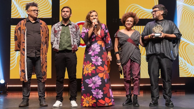 Cinco pessoas estão em um palco iluminado, em frente a um telão amarelo com padrões brancos e pretos. Da esquerda para a direita: um homem de óculos e cabelo grisalho curto usa camisa preta, calça jeans e um kimono marrom estampado; um homem de barba curta veste camisa verde, calça preta e jaqueta estampada; uma mulher no centro segura um microfone e usa vestido longo preto com grandes flores roxas, lilases, vermelhas, laranjas e amarelas; uma mulher com cabelo crespo curto veste blusa cinza transpassada com detalhe bordô, calça roxa e botas pretas; um homem segura um troféu dourado em forma de concha, veste blusa preta larga e calça preta.