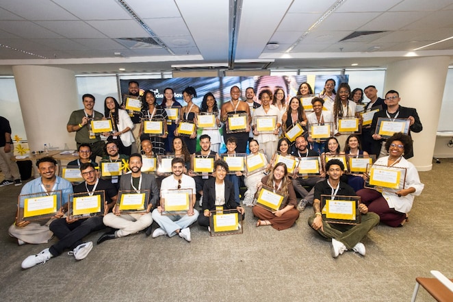 Foto de grupo com dezenas de pessoas reunidas em um ambiente corporativo, em pé e sentadas no chão, todas sorrindo e segurando certificados com moldura amarela. O espaço é uma sala ampla de escritório, com iluminação clara, colunas brancas e um telão ao fundo. As pessoas vestem roupas variadas, do casual ao social, e usam crachás no pescoço, sugerindo a conclusão ou celebração de um programa, treinamento ou reconhecimento coletivo.