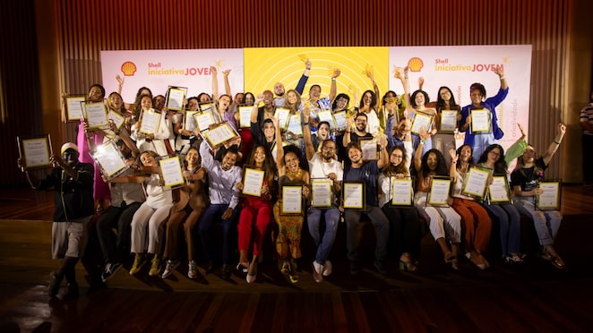 Grupo de pessoas sorrindo e segurando certificados, em celebração no palco de um evento do programa "Shell Iniciativa Jovem". Ao fundo, um painel com o logotipo da Shell e o nome do programa, decorado com tons amarelos e laranjas.